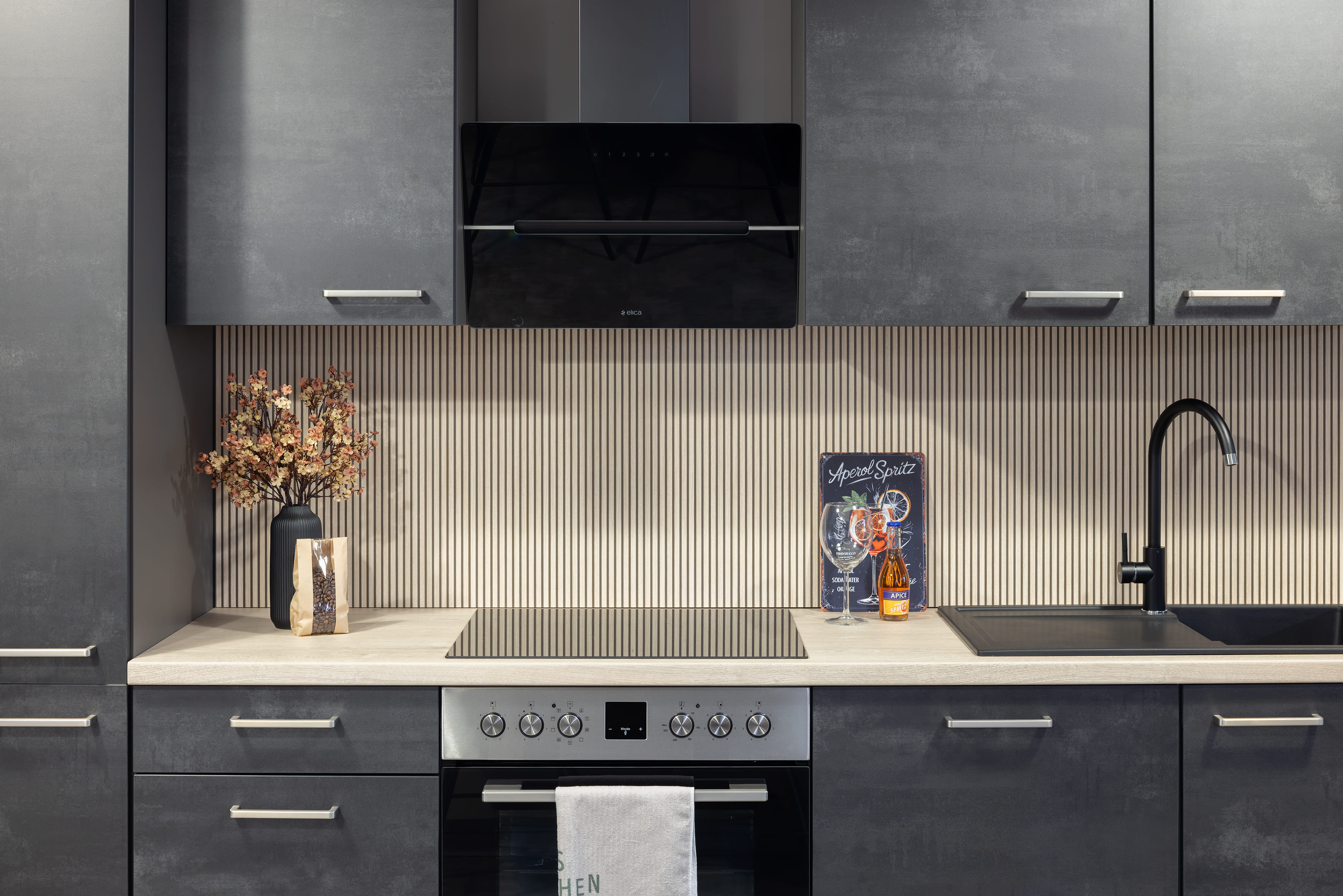 Moderne graue Küche mit Herd, Spüle, Vase und Kochbuch auf hellem Arbeitsbereich.