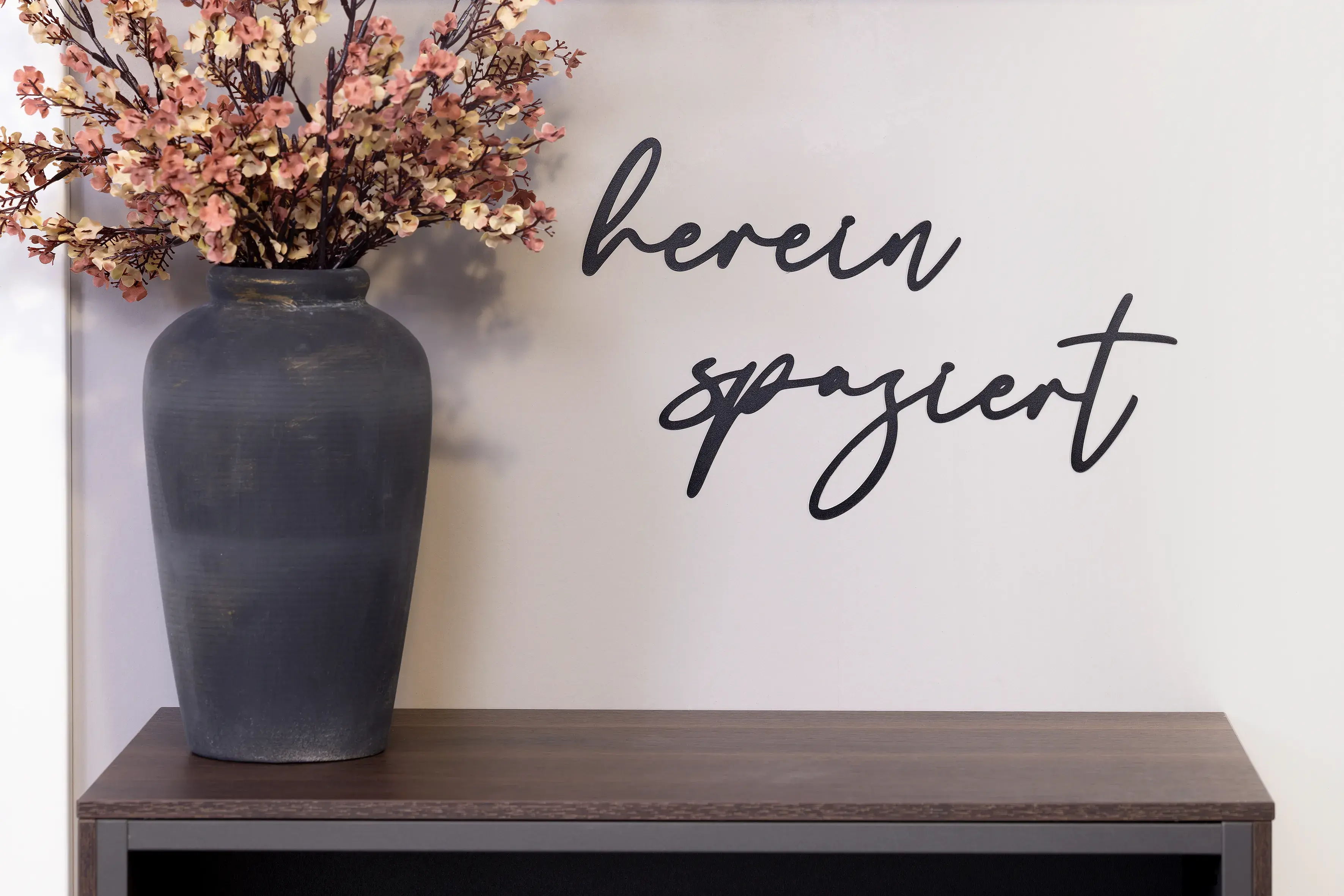 Graue Vase mit Trockenblumen auf Regal, Wand mit Schriftzug "herein spaziert"