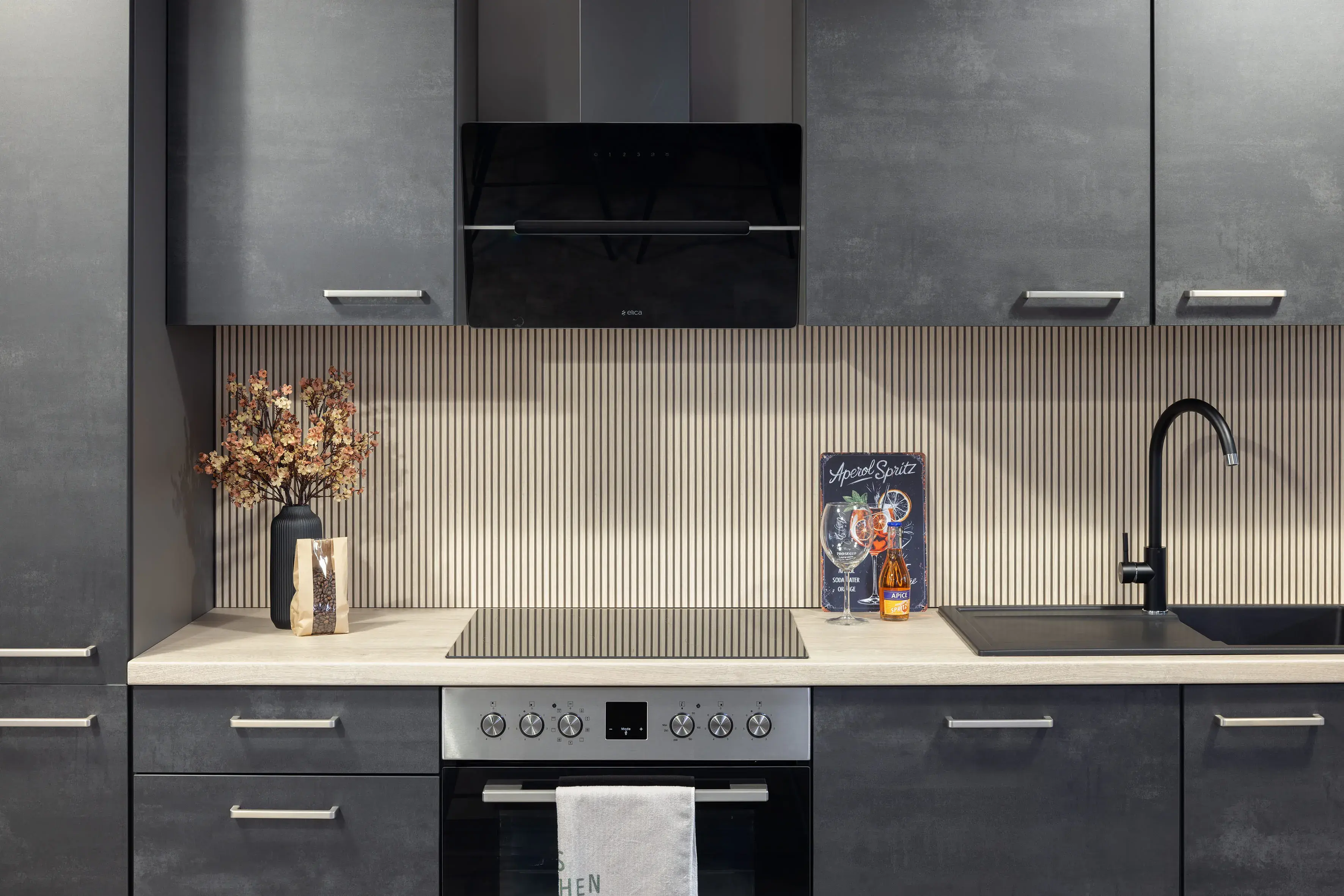 Moderne graue Küche mit Herd, Spüle, Vase und Kochbuch auf hellem Arbeitsbereich.